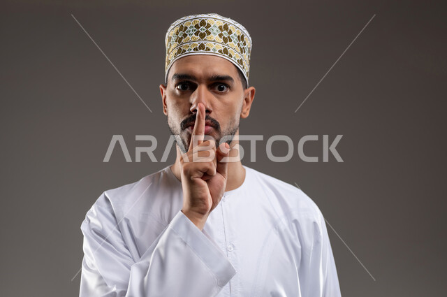مفهوم الحاجة الى التوقف والانصات، بورتريه لشاب عربي خليجي عماني يرتدي الدشداشة و الكمه يشير باصبع السبابه نحو فمه بايماءات الحاجه الى الهدوء والسكوت، حل المشكلات واتخاذ القرارات, إيماءات التمعن والتركيز والتخطيط للمستقبل، خلفيه رماديه