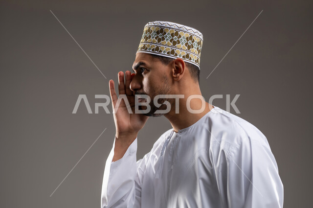 ايماءات التحذير والمناداه، بورتريه لرجل عربي خليجي عماني يرتدي الدشداشة والكمه يقوم بمناداة شخص ما بايماءات الانصات والتركيز، تعابير وجه ويد تدلان على التنبيه من حدوث مصيبة ما، خلفية رماديه