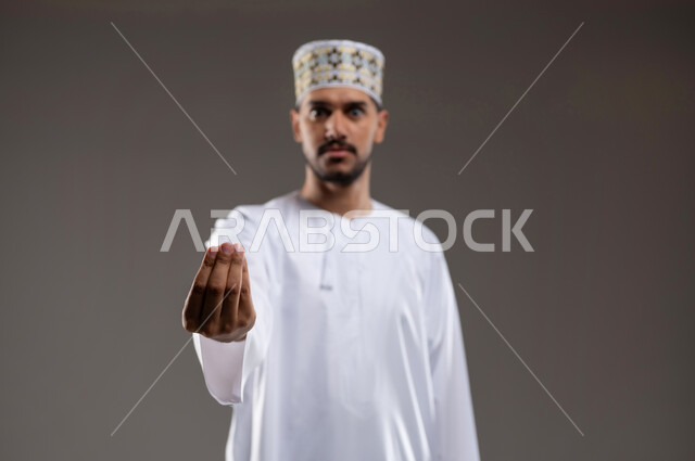 حركة يد تعني خفض الصوت والهدوء، ايماءات وجه ويدين تدل على محاولة تهدئة نقاش حاد، بورتريه لرجل عربي عماني يرتدي الدشداشة والكمه تظهر عليه علامات الانزعاج وعدم الراحة، طريقة الطلب بأدب واحترام، خلفية رماديه