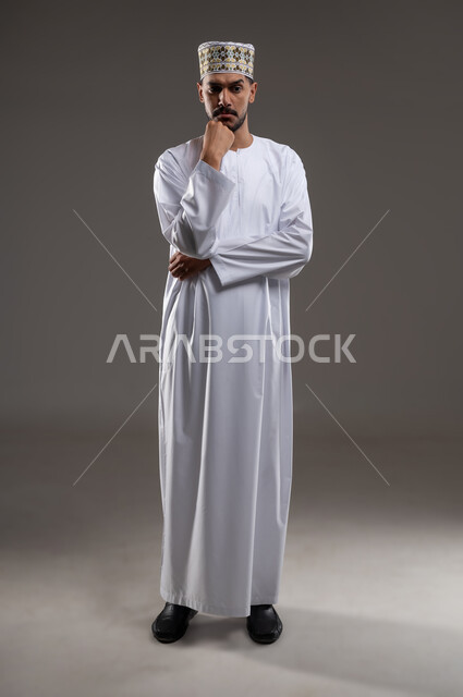 التفكير والتخطيط لشيء ما، ايماءات وجه تدل على التمعن والتدقيق، بورتريه لرجل عربي خليجي عماني يرتدي الدشداشة والكمه يشير بيده الى ذقنه، مفهوم النظرة المستقبلية، خلفية رماديه
