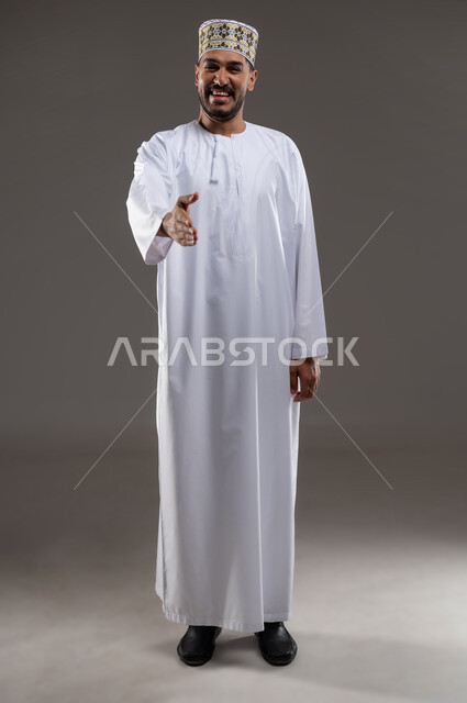 مد اليد للإمام اشارةً على التحيه والسلام، بورتريه لرجل عربي خليجي عماني يرتدي الدشداشة والكمه يمد يده بابتسامه  بايماءات الترحاب والمصافحه، خلفية رماديه