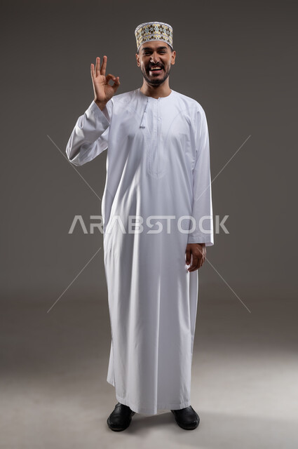 Gestures indicating approval and acceptance, portrait of an Arab Gulf Omani man wearing a dishdasha and a sleeve, pointing with the sign of quality, expressions of joy and happiness appear on him, gray background