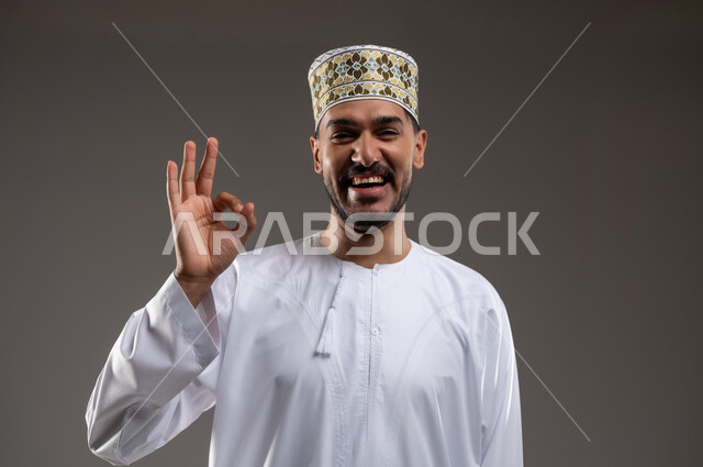 ايماءات تدل على الموافقة والقبول، بورتريه لرجل عربي خليجي عماني يرتدي الدشداشة والكمه يشير بعلامه الجوده تظهر عليه تعابير الفرح والسرور، خلفية رماديه