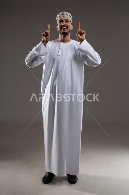 رفع اليدين للاعلى بايماءات القسم، بورتريه لشاب عربي خليجي عماني يرتدي الدشداشه والكمه يشير بأصبعي السبابه للاعلى بايماءات الشهاده وحلف اليمين، خلفيه رماديه