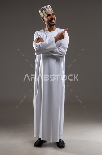 الإشارة والتوجيه الى مكان ما، بورتريه لرجل عربي خليجي عماني يرتدي الدشداشة والكمه يقف مبتسما ويشير باصبع السبابه الى مكان ما، تحديد موقع  او هدف معين، حركات واشارات، خلفيه رماديه