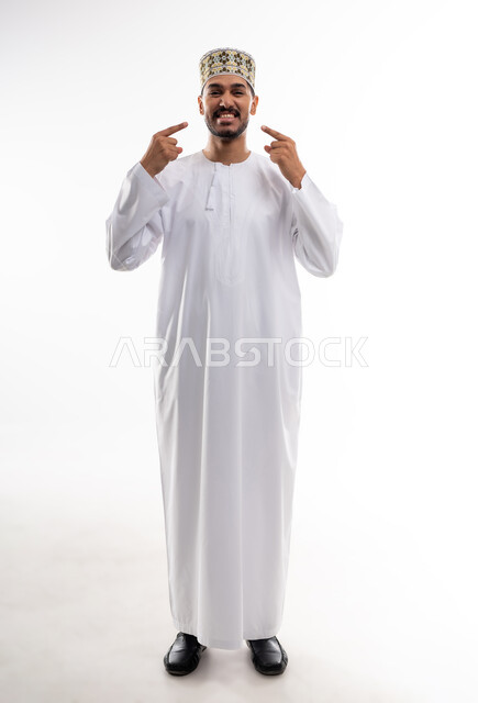 الاشارة إلى الفم بإيماءات الانزعاج والألم، مشاكل التهاب اللثة والحاجة لزيارة طبيب الأسنان, بورتريه  لشاب عربي خليجي عماني يرتدي الدشداشة و الكمه يشير الى فمه بايماءات الحاجه الى تلقي العلاج، خلفية بيضاء