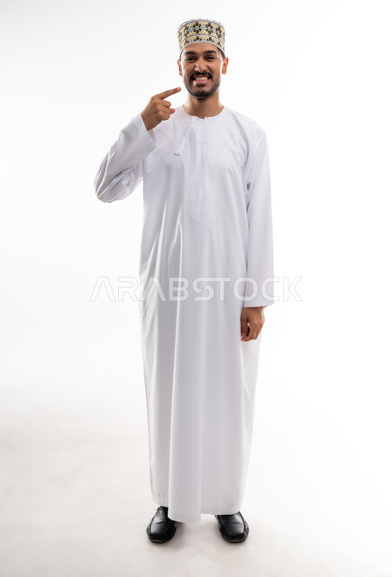 الاشارة إلى الفم بإيماءات الانزعاج والألم، مشاكل التهاب اللثة والحاجة لزيارة طبيب الأسنان, بورتريه  لشاب عربي خليجي عماني يرتدي الدشداشة و الكمه يشير الى فمه بايماءات الحاجه الى تلقي العلاج، خلفية بيضاء