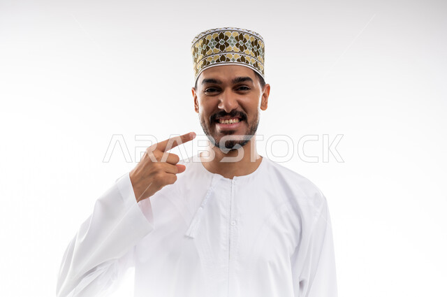 الاشارة إلى الفم بإيماءات الانزعاج والألم، مشاكل التهاب اللثة والحاجة لزيارة طبيب الأسنان, بورتريه  لشاب عربي خليجي عماني يرتدي الدشداشة و الكمه يشير الى فمه بايماءات الحاجه الى تلقي العلاج، خلفية بيضاء