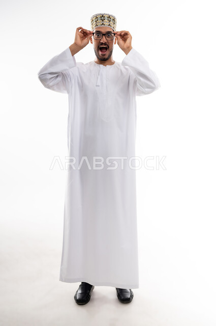 The concept of farsightedness and nearsightedness, feeling happy and joyful with clear vision, using eyeglasses to correct vision, portrait of an Arab Gulf Omani man wearing a dishdasha and a cloak holding his glasses with his hands with gestures of using visual aids using eyeglasses, white background