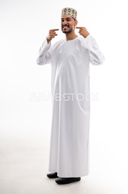 الاشارة إلى الفم بإيماءات الانزعاج والألم، مشاكل التهاب اللثة والحاجة لزيارة طبيب الأسنان, بورتريه  لشاب عربي خليجي عماني يرتدي الدشداشة و الكمه يشير الى فمه بايماءات الحاجه الى تلقي العلاج، خلفية بيضاء