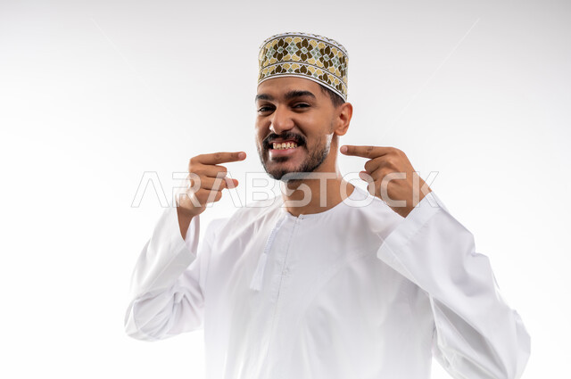 الاشارة إلى الفم بإيماءات الانزعاج والألم، مشاكل التهاب اللثة والحاجة لزيارة طبيب الأسنان, بورتريه  لشاب عربي خليجي عماني يرتدي الدشداشة و الكمه يشير الى فمه بايماءات الحاجه الى تلقي العلاج، خلفية بيضاء