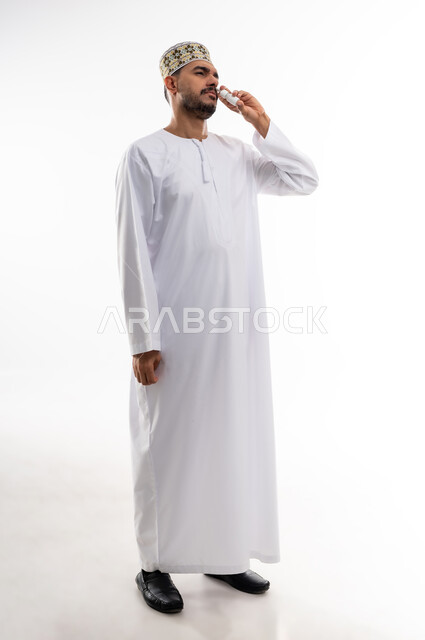 Treating chronic allergic rhinitis symptoms, maintaining a regular airway using nasal spray, portrait of an Arab Gulf Omani man wearing a dishdasha and a keffiyeh inhaling asthma and allergy medications, providing home health services, white background