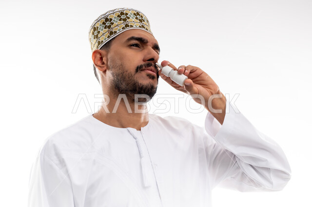 مداواة أعراض حساسيه الانف المزمنه، المحافظة على ممر هوائي منتظم باستخدام بخاخه الانف، بورتريه  لرجل عربي خليجي عماني يرتدي الدشداشة والكمه يقوم باستنشاق أدوية معالجة الربو والحساسيه، تقديم الخدمات الصحية المنزلية، خلفية بيضاء