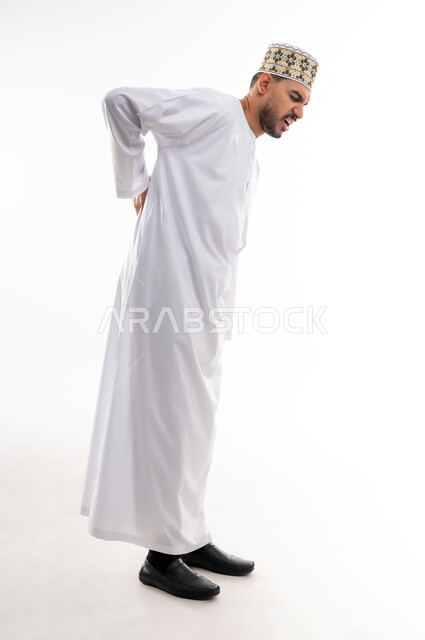 الأوجاع والآلام العصبية والعضلية، طلب المساعده والحاجه للعلاج، التهاب العضلات والمفاصل، بورتريه لرجل عربي خليجي عماني يرتدي الدشداشة و الكمه يضع يده على خصره بايماءات الألم والتعب والارهاق، حالة اسعافية طارئة, خلفية رماديه