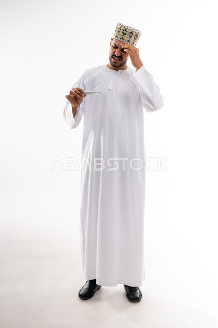 Symptoms of fever, physical fatigue and measuring body temperature, portrait of an Arab Gulf Omani man wearing a dishdasha and a sleeve reading his temperature using a digital thermometer, the concept of caring for health and the need for treatment, white background