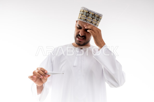 أعراض الاصابة بالحمى والتعب الجسدي وقياس درجه حراره الجسم،  بورتريه لرجل عربي خليجي عماني يرتدي الدشداشه والكمه يقوم بقراءه درجة حرارته باستخدام الميزان الحراري الرقمي, مفهوم الاهتمام بالصحة والحاجه الى العلاج, خلفية بيضاء