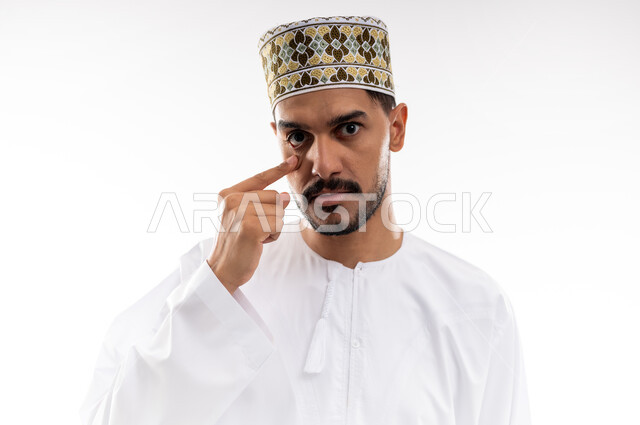 ارتفاع ضغط العين, الاحساس بالجفاف و احمرار العيون، الحاجة للفحص وعلاج حالات التهاب القرنيه، بورتريه لرجل عربي خليجي عماني يرتدي الدشداشه والكمه يشير باصبع السبابه نحو عينه بايماءات الحاجه الى العلاج وزياره الطبيب, خلفية بيضاء