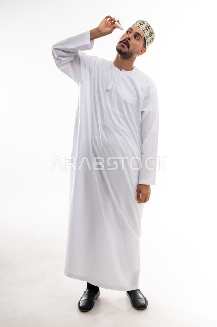 High eye pressure, feeling of dryness and redness of the eyes, need for examination and treatment of cases of corneal inflammation, portrait of an Arab Gulf Omani man wearing a dishdasha and a cuff putting in eye drops, white background