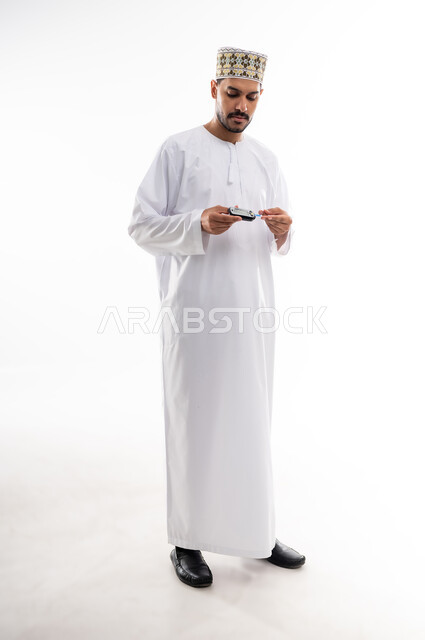 الالام الجسدية والحالات المرضية, مشاكل ارتفاع السكر في الدم, بورتريه لرجل عربي خليجي عماني يرتدي الدشداشه والكمه يقوم بفحص نسبة السكر في الدم باستخدام جهاز الفحص السريع, الأجهزة الالكترونية الحديثة لمرضى داء السكري, خلفية بيضاء