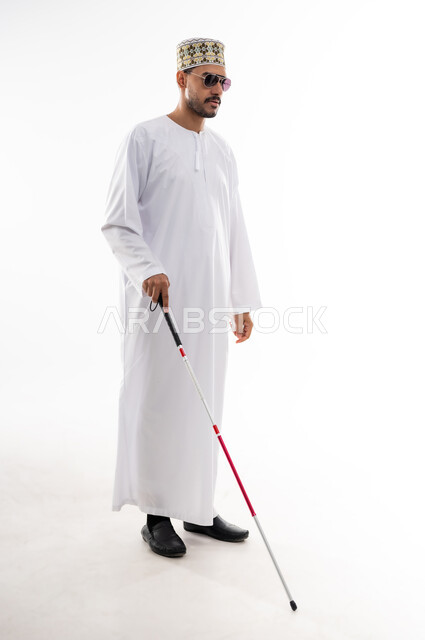 Poor visual ability, gestures of pleasure and happiness, portrait of a blind Arab Gulf Omani man wearing a dishdasha and a sleeve and putting sunglasses on his eyes, holding a stick in his hand to help him move to the right place, white background