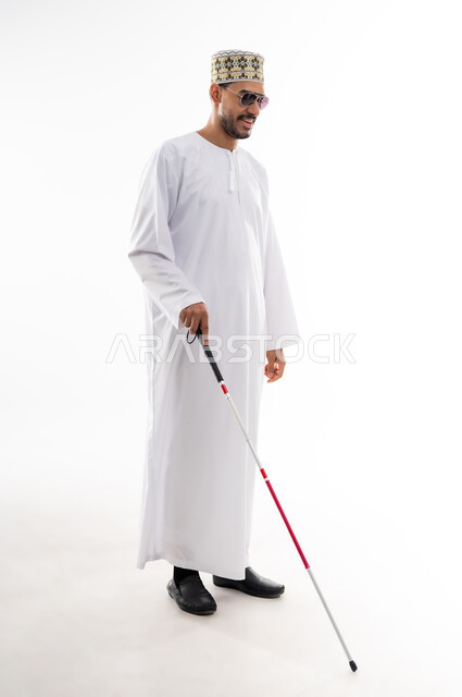 ضعف القدرة البصرية، ايماءات السرور والسعادة، بورتريه  لرجل عربي خليجي عماني كفيف يرتدي الدشداشه والكمه ويضع النظارة الشمسية على عينه يمسك بيده عصا لمساعدته على الحركه الى المكان الصحيح، خلفيه بيضاء