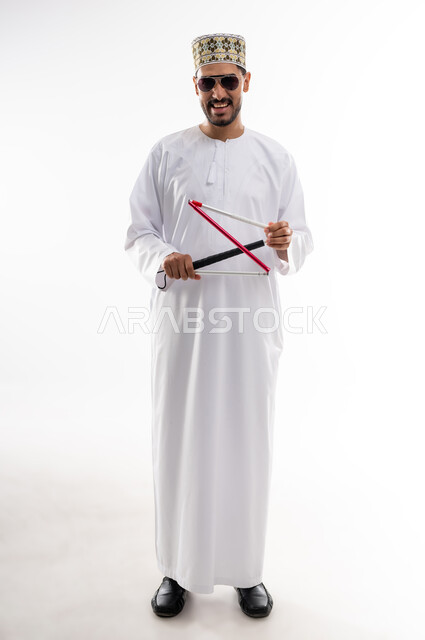ضعف القدرة البصرية، ايماءات السرور والسعادة، بورتريه  لرجل عربي خليجي عماني كفيف يرتدي الدشداشه والكمه ويضع النظارة الشمسية على عينه يمسك بيده عصا لمساعدته على الحركه الى المكان الصحيح، خلفيه بيضاء