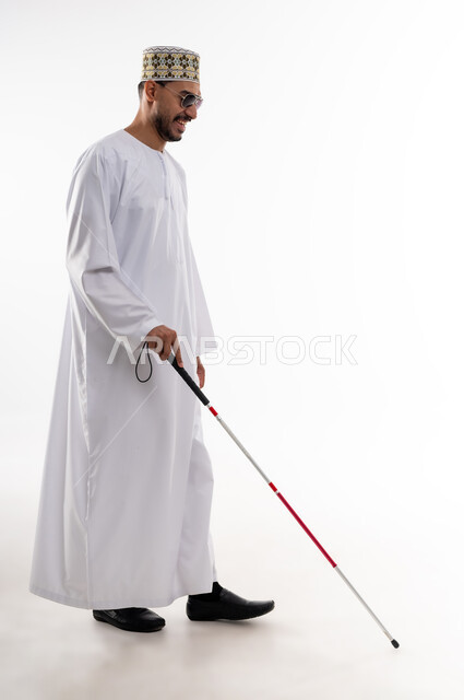ضعف القدرة البصرية، ايماءات السرور والسعادة، بورتريه  لرجل عربي خليجي عماني كفيف يرتدي الدشداشه والكمه ويضع النظارة الشمسية على عينه يمسك بيده عصا لمساعدته على الحركه الى المكان الصحيح، خلفيه بيضاء
