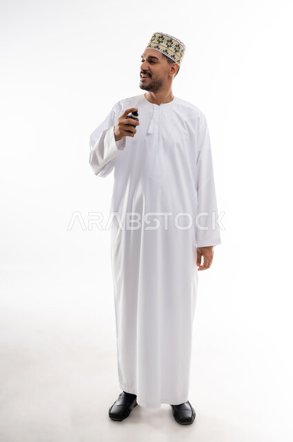 استخدام الزيوت العطرية الطبيعية, عطورات عربيه خليجيه, مفهوم الاناقة والاهتمام بالمظهر الخارجي, بورتريه  لرجل عربي خليجي عماني يرتدي الدشداشه والكمه يقوم برش العطر على ثوبه, التسويق لمنتجات العطور, خلفيه بيضاء