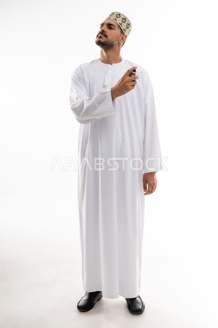 استخدام الزيوت العطرية الطبيعية, عطورات عربيه خليجيه, مفهوم الاناقة والاهتمام بالمظهر الخارجي, بورتريه  لرجل عربي خليجي عماني يرتدي الدشداشه والكمه يقوم برش العطر على ثوبه, التسويق لمنتجات العطور, خلفيه بيضاء