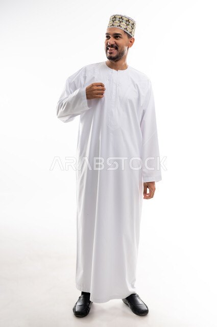 Gestures of pleasure and self-confidence, the concept of elegance and masculinity, taking personal photos for official documents, a portrait of a young Arab Gulf Omani man wearing a dishdasha and a turban, standing straight, looking at the camera with a smile, taking care and attention to one’s external appearance, white background.