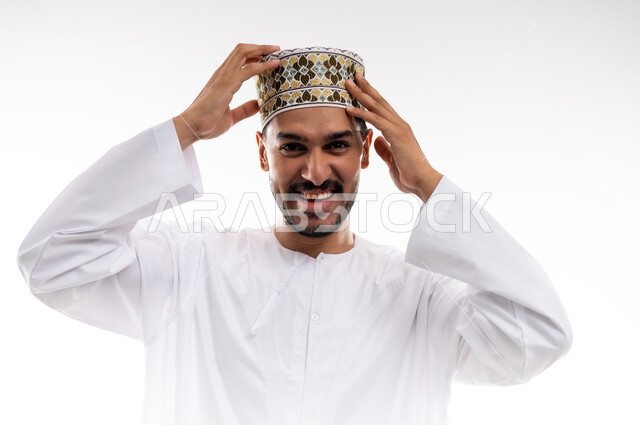 إيماءات السرور والثقة بالنفس، مفهوم الاناقة والرجولة، التقاط صور شخصية للوثائق الرسمية، بورتريه لشاب عربي خليجي عماني يرتدي الدشداشه والكمه يقف باستقامة ينظر الى الكاميرا بابتسامه، الاعتناء والاهتمام بالمظهر الخارجي، خلفية بيضاء .