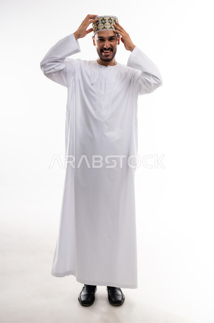 Gestures of pleasure and self-confidence, the concept of elegance and masculinity, taking personal photos for official documents, a portrait of a young Arab Gulf Omani man wearing a dishdasha and a turban, standing straight, looking at the camera with a smile, taking care and attention to one’s external appearance, white background.