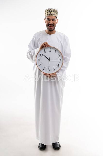 The concept of the importance of time and commitment to appointments, a portrait of an Arab Gulf Omani man wearing a dishdasha and a cuff, holding a large digital watch and pointing his thumb upwards in gestures of commitment to time, completing tasks and work on time, white background