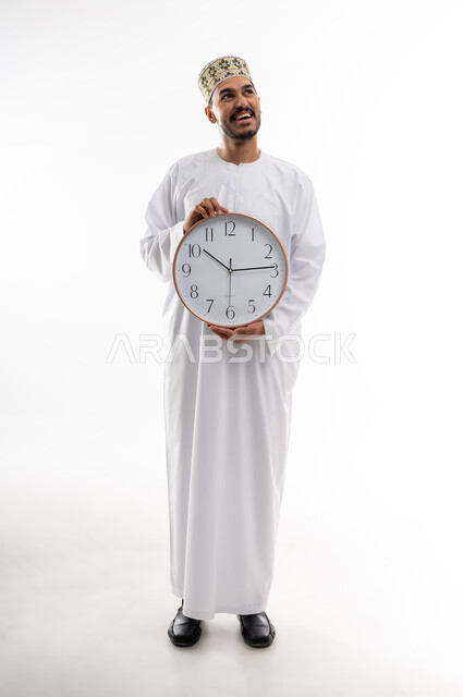 مفهوم اهميه الوقت والالتزام بالمواعيد، بورتريه لرجل عربي خليجي عماني يرتدي الدشداشه والكمه  يحمل ساعة رقمية كبيرة ويشير باصبع الابهام نحو الاعلى بايماءات الالتزام بالوقت، إنجاز المهامات والاعمال على الوقت، خلفيه بيضاء