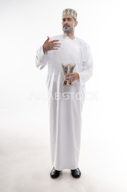 استخدام خشب العود بالأعياد والمناسبات, التطيب بالعطور والبخور, بورتريه لشاب عربي خليجي عماني يرتدي الدشداشه والكمه يحمل بيده المبخرة ويقوم بالتعطر, الأناقة والاهتمام بالمظهر الخارجي, العادات والتقاليد العمانيه, خلفيه بيضاء