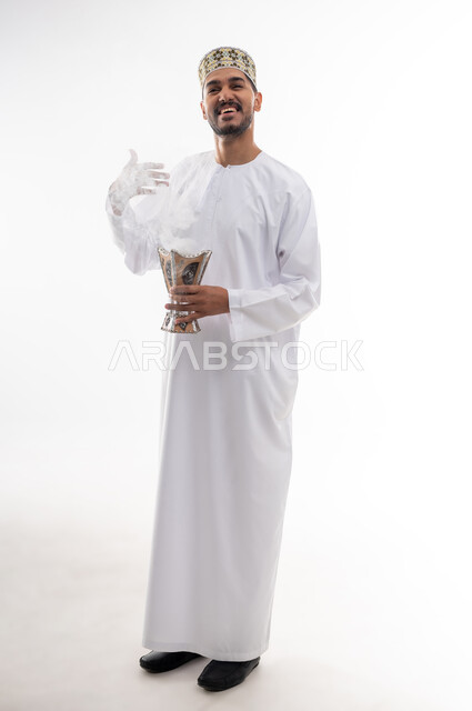 استخدام خشب العود بالأعياد والمناسبات, التطيب بالعطور والبخور, بورتريه لشاب عربي خليجي عماني يرتدي الدشداشه والكمه يحمل بيده المبخرة ويقوم بالتعطر, الأناقة والاهتمام بالمظهر الخارجي, العادات والتقاليد العمانيه, خلفيه بيضاء