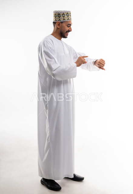 The concept of the importance of time and commitment to appointments, a portrait of an Arab Gulf Omani man wearing a dishdasha and a kummah looking at his personal watch with gestures of the importance of commitment to time and completing tasks and work accurately, white background