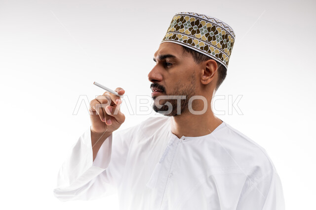 الادمان على مشتقات التبغ والسجائر، عادات سلوكيه ضاره بالصحه وتعاطي المواد السامه،  بورتريه لرجل عربي خليجي عماني يرتدي الدشداشه والكمه يقوم بتدخين السجائر، خلفية بيضاء