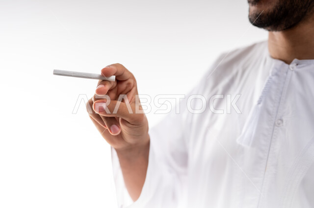 Addiction to tobacco and cigarette derivatives, harmful behavioral habits and use of toxic substances, portrait of an Arab Gulf Omani man wearing a dishdasha and a kummah smoking cigarettes, white background