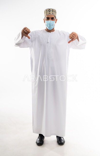 Rejection, non-acceptance and satisfaction, avoiding the spread of infection from Covid-19, maintaining public safety and following precautionary measures to prevent the Corona virus, portrait of a young Arab Gulf Omani man wearing a dishdasha and a kuma and putting on a medical mask, pointing his thumbs down, white background