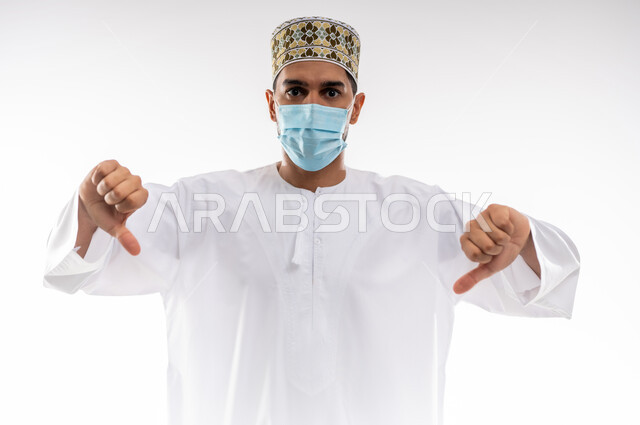 Rejection, non-acceptance and satisfaction, avoiding the spread of infection from Covid-19, maintaining public safety and following precautionary measures to prevent the Corona virus, portrait of a young Arab Gulf Omani man wearing a dishdasha and a kuma and putting on a medical mask, pointing his thumbs down, white background