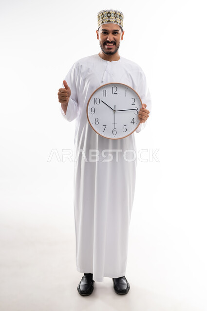 مفهوم اهميه الوقت والالتزام بالمواعيد، بورتريه لرجل عربي خليجي عماني يرتدي الدشداشه والكمه  يحمل ساعة رقمية كبيرة ويشير باصبع الابهام نحو الاعلى بايماءات الالتزام بالوقت، إنجاز المهامات والاعمال على الوقت، خلفيه بيضاء
