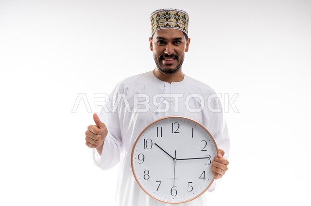 مفهوم اهميه الوقت والالتزام بالمواعيد، بورتريه لرجل عربي خليجي عماني يرتدي الدشداشه والكمه  يحمل ساعة رقمية كبيرة ويشير باصبع الابهام نحو الاعلى بايماءات الالتزام بالوقت، إنجاز المهامات والاعمال على الوقت، خلفيه بيضاء