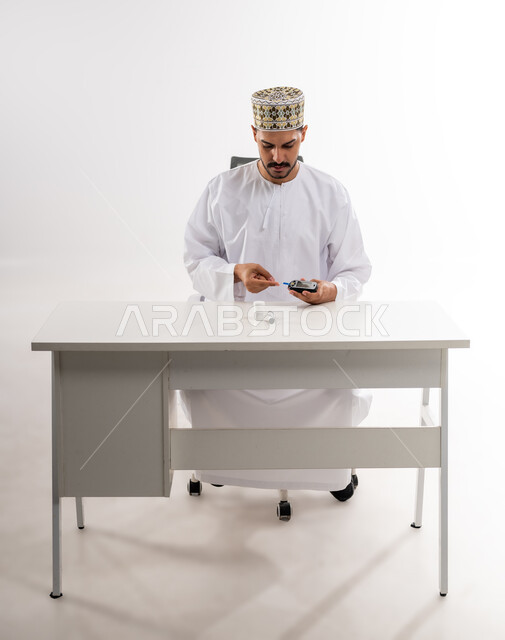 الالام الجسدية والحالات المرضية, مشاكل ارتفاع السكر في الدم, بورتريه لرجل عربي خليجي عماني يرتدي الدشداشه والكمه يقوم بفحص نسبة السكر في الدم باستخدام جهاز الفحص السريع, الأجهزة الالكترونية الحديثة لمرضى داء السكري, خلفية بيضاء