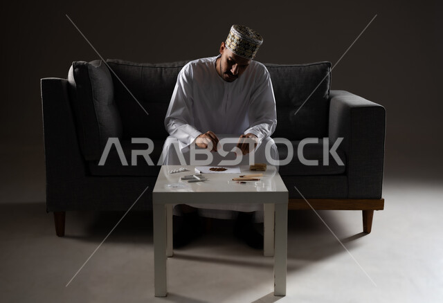 القيام بعادات سلوكية ضارة وتعاطي المواد السامة، الأثار السلبية للادمان على التبغ والمخدرات، المعاناة من الاضطرابات النفسية والمشاكل الصحية، بورتريه لرجل عربي خليجي عماني يرتدي الدشداشه والكمه يجلس على اريكة ويقوم بلف سجائر الحشيش المخدر، صوره ظليه