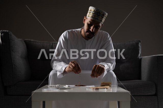 القيام بعادات سلوكية ضارة وتعاطي المواد السامة، الأثار السلبية للادمان على التبغ والمخدرات، المعاناة من الاضطرابات النفسية والمشاكل الصحية، بورتريه لرجل عربي خليجي عماني يرتدي الدشداشه والكمه يجلس على اريكة ويقوم بلف سجائر الحشيش المخدر، صوره ظليه