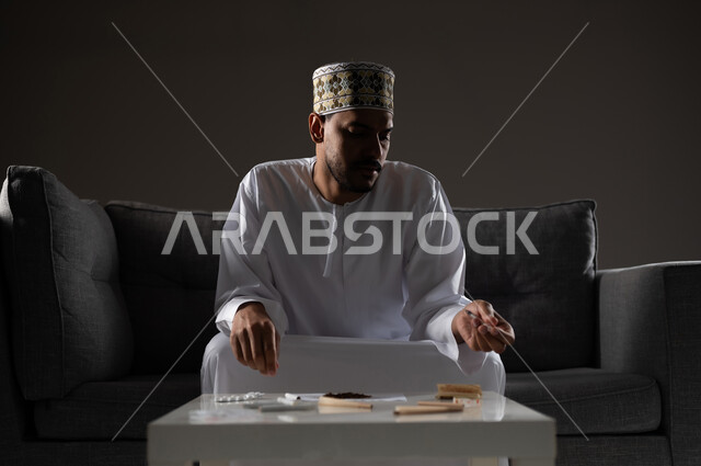 القيام بعادات سلوكية ضارة وتعاطي المواد السامة، الأثار السلبية للادمان على التبغ والمخدرات، المعاناة من الاضطرابات النفسية والمشاكل الصحية، بورتريه لرجل عربي خليجي عماني يرتدي الدشداشه والكمه يجلس على اريكة ويقوم بلف سجائر الحشيش المخدر، صوره ظليه