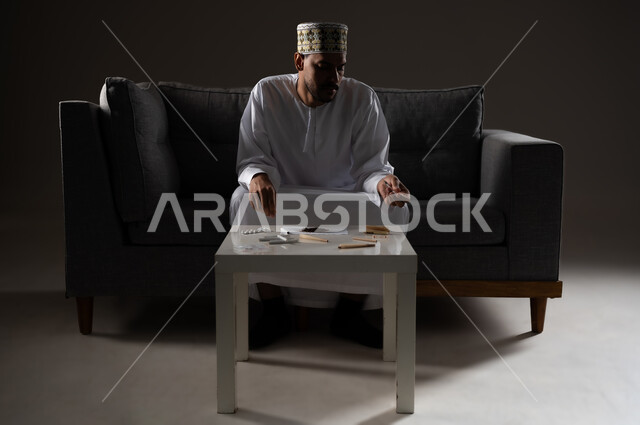 القيام بعادات سلوكية ضارة وتعاطي المواد السامة، الأثار السلبية للادمان على التبغ والمخدرات، المعاناة من الاضطرابات النفسية والمشاكل الصحية، بورتريه لرجل عربي خليجي عماني يرتدي الدشداشه والكمه يجلس على اريكة ويقوم بلف سجائر الحشيش المخدر، صوره ظليه