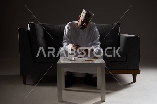 القيام بعادات سلوكية ضارة وتعاطي المواد السامة، الأثار السلبية للادمان على التبغ والمخدرات، المعاناة من الاضطرابات النفسية والمشاكل الصحية، بورتريه لرجل عربي خليجي عماني يرتدي الدشداشه والكمه يجلس على اريكة ويقوم بلف سجائر الحشيش المخدر، صوره ظليه
