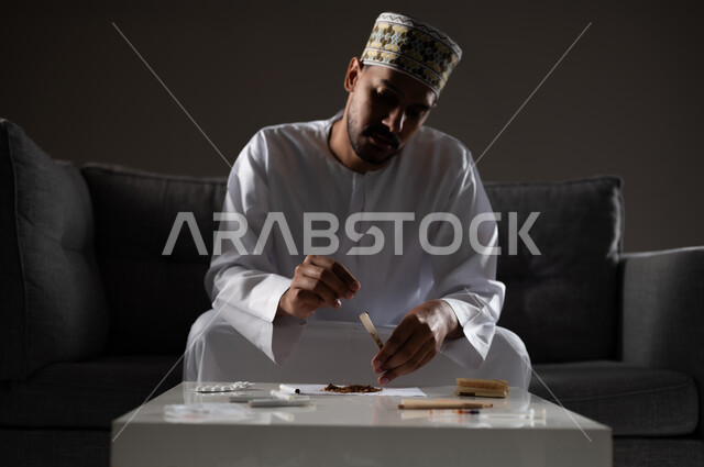 القيام بعادات سلوكية ضارة وتعاطي المواد السامة، الأثار السلبية للادمان على التبغ والمخدرات، المعاناة من الاضطرابات النفسية والمشاكل الصحية، بورتريه لرجل عربي خليجي عماني يرتدي الدشداشه والكمه يجلس على اريكة ويقوم بلف سجائر الحشيش المخدر، صوره ظليه